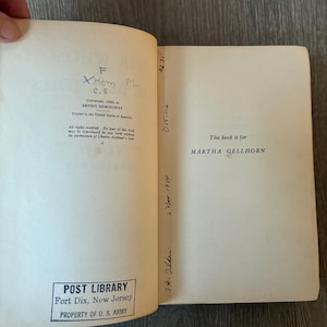 以下が含まれることがあります： 「ERNEST HEMINGWAY」と「MARTHA GELLHORN」を含むテキストが書かれた開いた本。本には、ニュージャージー州フォートディックスの郵便図書館のスタンプと手書きのメモと数字が記載されています。