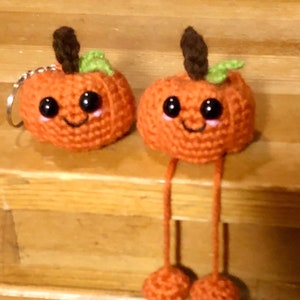 Op de afbeelding: Twee oranje gehaakte pompoenen met lachende gezichten. Eén pompoen heeft een sleutelhanger. De pompoenen hebben bruine stelen en groene bladeren. Eén pompoen heeft lange oranje benen met oranje voeten.