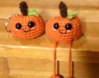 May include: Two orange crochet pumpkins with smiling faces. One pumpkin has a keychain attached. The pumpkins have brown stems and green leaves. One pumpkin has long orange legs with orange feet.
