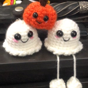 Op de afbeelding: Een trio handgemaakte Halloween-decoraties van gehaakt garen. De set bevat twee witte spoken met bungelende benen en een lachende oranje pompoen. Elke figuur heeft zwarte ogen en roze wangen.