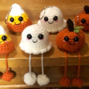 May include: A collection of handmade crochet Halloween-themed keychains. The keychains include smiling candy corn, ghosts, and pumpkins, each with dangling legs. The candy corn is orange, yellow, and white. The ghosts are white, and the pumpkins are orange.