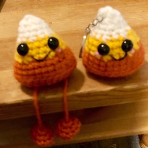 Op de afbeelding: Twee handgemaakte gehaakte snoepmaïsvormen. Elke snoepmaïs heeft een witte bovenkant, een gele kern en een oranje basis. Ze hebben zwarte ogen, roze wangen en lachende monden. Eén heeft bungelende oranje benen met oranje pompons, de andere heeft een sleutelhanger.