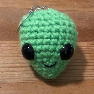 May include: A handmade, green crocheted alien head with large, black, glossy eyes and a stitched smile. The alien has a silver keychain attached to the top. The background is a wooden surface.