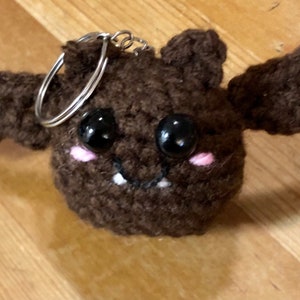 May include: A handmade brown crochet bat keychain with black bead eyes, pink cheeks, and small white fangs. The bat has outstretched wings and a silver keyring attached to its head. This whimsical accessory is perfect for Halloween or as a unique gift.