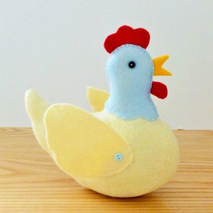 May include: A yellow felt chicken with a blue head, red comb, and yellow beak. The chicken has a button on its wing.