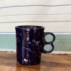 May include: A white ceramic mug with a black handle and the text "But First, Coffee" in a black, handwritten-style font. The mug is on a white surface.