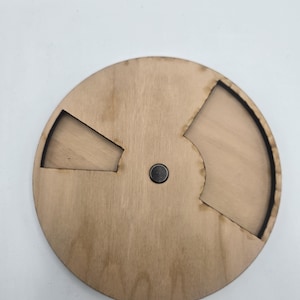 May include: A round wooden puzzle with two cut-out sections and a small metal knob in the center. The puzzle is made of light-colored wood and has a simple, geometric design. The cut-out sections are in the shape of a triangle and a curved section.