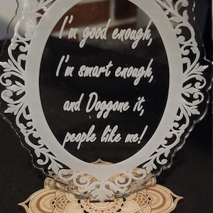 May include: A white acrylic framed sign with a decorative wooden base. The sign reads "I'm good enough, I'm smart enough, and Doggone it, people like me!"