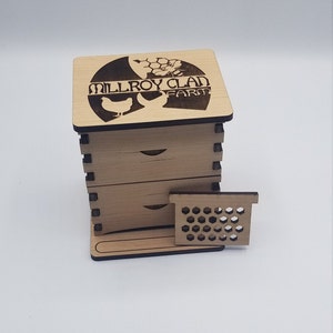 May include: A wooden box with three drawers and a honeycomb-shaped vent. The box is labeled "Millroy Clan Farm" with a bee and flower design.