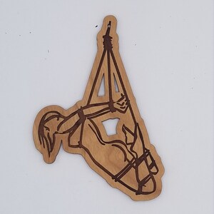 Op de afbeelding: Een houten ornament dat een persoon voorstelt die met touw vastgebonden is in een hangende positie. Het ornament is gesneden uit lichtbruin hout en heeft een donkerbruine vlek.