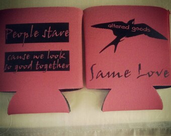 May include: Two red can coolers with black text and graphics. The left cooler says "People stare cause we look so good together". The right cooler says "altered goods" and has a black silhouette of a bird. The right cooler also says "Same Love".