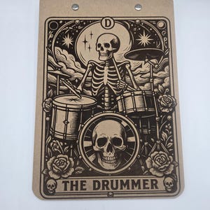May include: A brown clipboard with a black and white illustration of a skeleton drummer. The design includes a skull, roses, and the text "THE DRUMMER". The clipboard has a metal clip at the top.