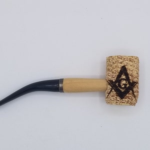 May include: A wooden corn cob pipe with a black stem. The bowl of the pipe is decorated with a black and white image of a compass and square, a symbol of Freemasonry.