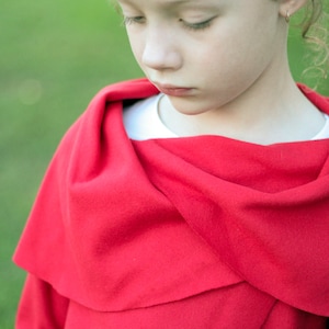 May include: A bright red cape draped over a white shirt. The cape has a wide, flowing design, with a soft, textured appearance. The background is a blurred green, suggesting an outdoor setting. The cape is the focal point.
