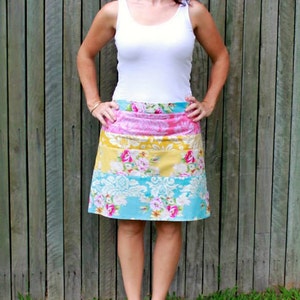 以下が含まれることがあります： 白いタンクトップとカラフルな花柄のスカートを着用した女性。スカートはピンク、イエロー、ブルー、ホワイトの柄で、花柄のデザインが施されています。彼女は木製のフェンスの前に立っています。