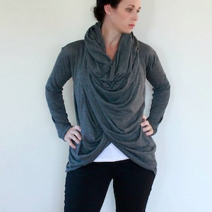 May include: A grey long-sleeved shirt with a cowl neck and a criss-cross design at the front. The shirt is worn over a white t-shirt and black trousers.