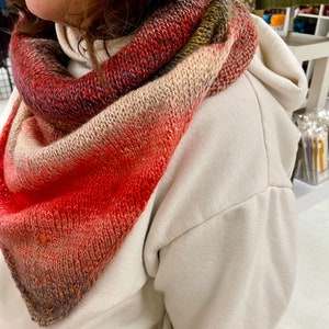 May include: A triangular hand-knitted shawl, displaying a colour gradient from deep red to beige. The shawl is wrapped around the neck, highlighting the textured knit and colour transitions.