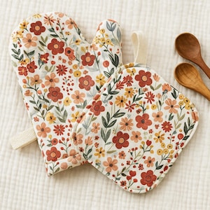 Peut inclure: Ensemble de manique et de dessous de plat à motifs floraux. Le tissu blanc est orné de fleurs rouges, oranges et jaunes avec des feuilles vertes. Une paire de cuillères en bois est en arrière-plan.