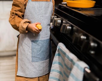 以下が含まれることがあります： 茶色のシャツを着て、青と白のストライプのエプロンをした若い人が、黒いストーブの前に立っています。彼らは右手に黄色いおもちゃを持っています。青と白のストライプのタオルがストーブにかけられています。