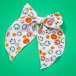 Op de afbeelding: Witte stoffen haarband met een patroon van kleurrijke smiley-gezichten. De strik is in een knoop geknoopt en heeft een witte achtergrond.