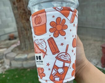 May include: A white and orange coffee cup sleeve with a fall-themed pattern. The design includes pumpkins, flowers, and coffee drinks. The sleeve is wrapped around a clear plastic cup with a lid.