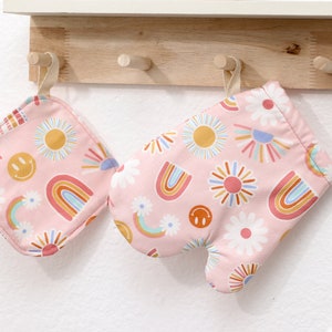 May include: A set of two pink oven mitts and a pot holder with a cheerful pattern of rainbows, suns, and daisies. The pattern is in shades of pink, orange, yellow, blue, and green.