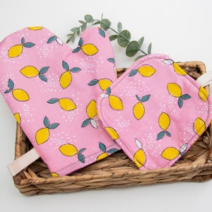 May include: A pink oven mitt and pot holder set with a lemon print. The set is made of cotton and has a white trim. The mitt has a loop for hanging.