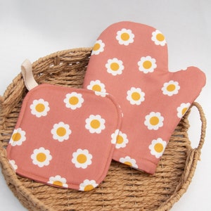 May include: A matching oven mitt and pot holder set. The set features a repeating pattern of white daisies with yellow centers on a dusty rose background. The pot holder has a square shape, and the oven mitt has a loop for hanging.