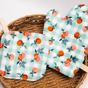 May include: A set of two kitchen pot holders with a green and white gingham pattern and a peach design. The pot holders are in a wicker basket.