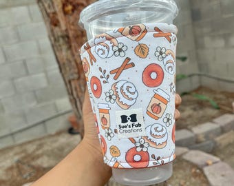 May include: A white coffee cup sleeve with a fall-themed pattern featuring donuts, cinnamon sticks, pumpkins, and flowers. The sleeve has a label that reads "Sue's Fab Creations". The sleeve is wrapped around a clear plastic cup.