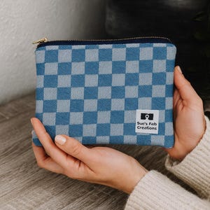May include: A blue and gray checkered pattern zippered pouch with a gold zipper. The pouch has a small white label with the text "Sue's Fab Creations" and a camera icon.