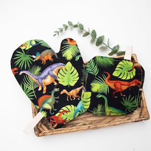 Puede incluir: Un par de guantes para horno negros con un patrón de dinosaurio y hojas tropicales. Los guantes están hechos de tela y tienen un lazo para colgar.