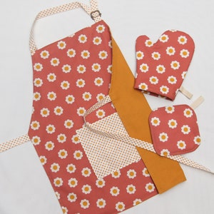 May include: A red and white floral print kitchen apron with a yellow and white polka dot pocket and matching oven mitts and pot holder.