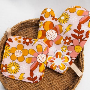 Op de afbeelding: Een roze ovenwant en pannenlap set met een retro bloemenpatroon in oranje, geel en bruin. Het ontwerp bevat een lachende bloem en een gele bliksemschicht. De set is geplaatst in een gevlochten mand.