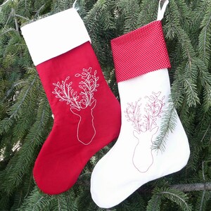 May include: Two red and white Christmas stockings with embroidered deer and foliage designs. The stocking on the left is red with a white cuff and the stocking on the right is white with a red polka dot cuff.