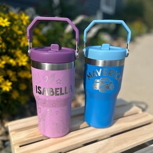 May include: Two stainless steel tumblers with handles. One is purple with the name "ISABELLA" and decorative etched designs. The other is blue with "MAVERICK" and a monster truck design. Both have matching colored lids and handles.