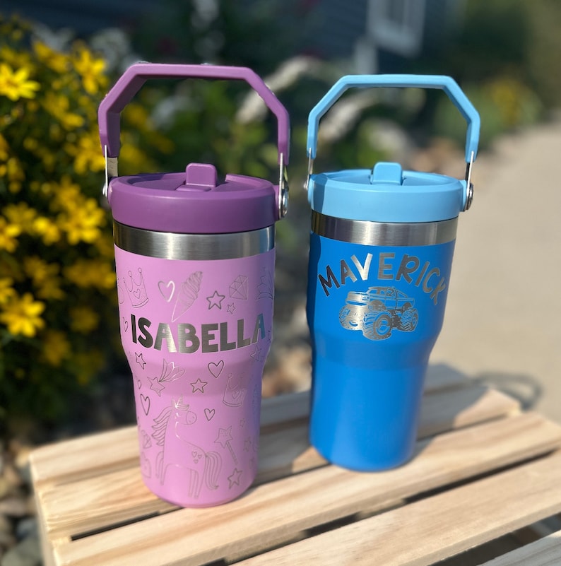 May include: Two stainless steel tumblers with handles. One is purple with the name "ISABELLA" and decorative etched designs. The other is blue with "MAVERICK" and a monster truck design. Both have matching colored lids and handles.