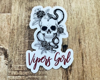 May include: A white skull with a black snake wrapped around it, with black roses and leaves. The text "Vipers Girl" is written in red below the skull.