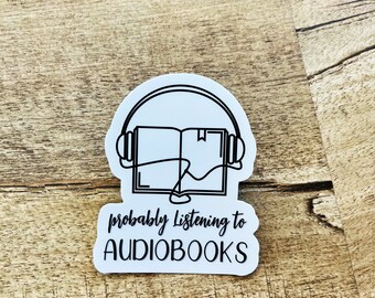 Op de afbeelding: Witte sticker met een zwarte omtrek van een koptelefoon met een boek erin. De tekst "probably listening to audiobooks" staat onder de afbeelding.