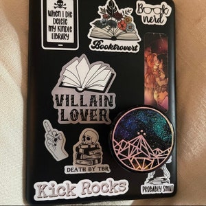 K&ouml;nnte beinhalten: Ein schwarzer Laptop mit verschiedenen Aufklebern, darunter ein Buch mit dem Text "Booktrovert", ein Sch&auml;del mit dem Text "When I die delete my kindle library", ein Buch mit dem Text "Villain Lover", eine Hand, die den Mittelfinger zeigt, ein Sch&auml;del auf einem Stapel B&uuml;cher mit dem Text "Death by TBR", ein Aufkleber mit dem Text "Kick Rocks", ein Aufkleber mit dem Text "Probably Smut" und ein runder Aufkleber mit einem Galaxie-Design und einer Bergkette.