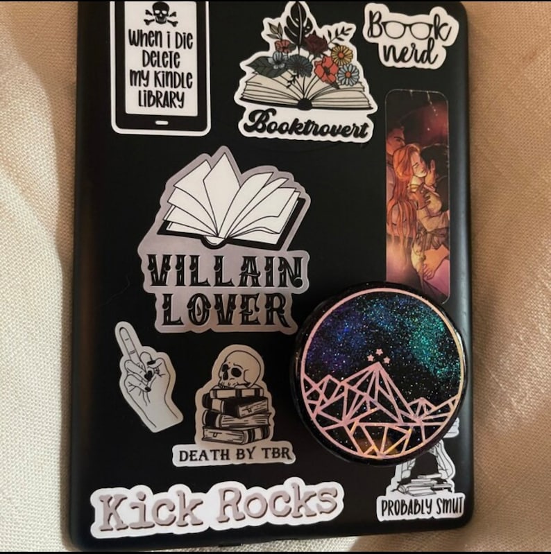 K&ouml;nnte beinhalten: Ein schwarzer Laptop mit verschiedenen Aufklebern, darunter ein Buch mit dem Text "Booktrovert", ein Sch&auml;del mit dem Text "When I die delete my kindle library", ein Buch mit dem Text "Villain Lover", eine Hand, die den Mittelfinger zeigt, ein Sch&auml;del auf einem Stapel B&uuml;cher mit dem Text "Death by TBR", ein Aufkleber mit dem Text "Kick Rocks", ein Aufkleber mit dem Text "Probably Smut" und ein runder Aufkleber mit einem Galaxie-Design und einer Bergkette.
