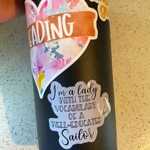 Op de afbeelding: Een zwarte herbruikbare waterfles met verschillende stickers. De stickers bevatten een hart met het woord "READING" erop, een sticker met de tekst "I'm a lady with the vocabulary of a well-educated sailor" en een sticker met de tekst "Kind go".