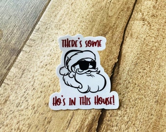 May include: A white sticker with a cartoon Santa Claus wearing sunglasses and a beard. The text "THERE'S SOME HO'S IN THIS HOUSE!" is written in red. The sticker is placed on a wooden surface.