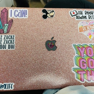 Op de afbeelding: Een roze laptop met verschillende stickers, waaronder een Mickey Mouse-sticker, een sticker met de tekst "The book is always better", een sticker met de tekst "Reading", een sticker met de tekst "You got this", een sticker met de tekst "I can!" en een sticker met de tekst "Zicke Zacke Zicke Zacke Hoi Hoi Hoi".