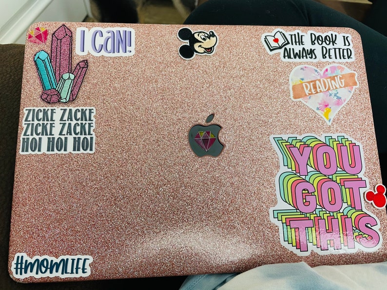 Op de afbeelding: Een roze laptop met verschillende stickers, waaronder een Mickey Mouse-sticker, een sticker met de tekst "The book is always better", een sticker met de tekst "Reading", een sticker met de tekst "You got this", een sticker met de tekst "I can!" en een sticker met de tekst "Zicke Zacke Zicke Zacke Hoi Hoi Hoi".