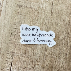 Puede incluir: Una pegatina blanca con texto negro que dice "Me gustan mis novios de libros oscuros y melancólicos". La pegatina está sobre una superficie de madera marrón claro.