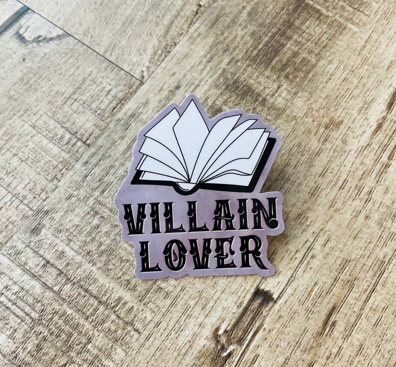 Op de afbeelding: Een zilveren sticker met een zwart-witte illustratie van een open boek en de tekst "VILLAIN LOVER" in zwart.