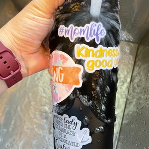 Op de afbeelding: Zwarte herbruikbare waterfles met verschillende kleurrijke stickers. De stickers zeggen "#momlife", "Vriendelijkheid is goed", en "Ik ben een dame met de woordenschat van een goed opgeleide zeeman".