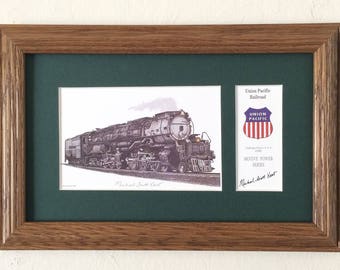 May include: A framed print of a black and white illustration of a Union Pacific Railroad train, the "Challenger" locomotive, with the text "Challenger Steam Train" below the image. The print is accompanied by a Union Pacific Railroad logo and a signature by Michael Albert Hart.