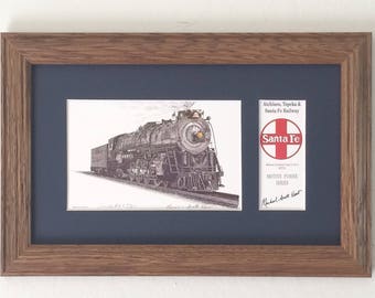 May include: A framed black and white illustration of a Santa Fe steam locomotive with the text "Atchison, Topeka & Santa Fe Railway" and "Motive Power Series" printed on a white card with a red cross and the words "Santa Fe" in a red circle. The card is signed "Michael Hart Hart".